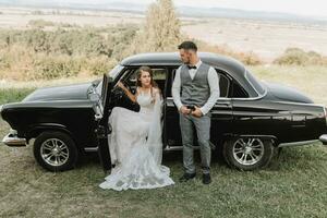 Side view of married bride and groom dressed in festive clothes standing against black retro car on their wedding day photo