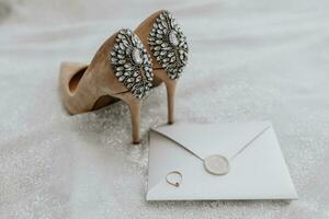 wedding shoes of the bride on a high heel of beige color with stones above the heel, an envelope with a wedding vow and a wedding ring of the bride is standing next to it photo