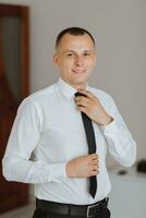 handsome smiling and stylish groom in a white shirt with a tie in his hands, preparing for the wedding photo