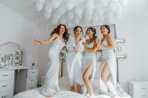 joven damas de honor en blanco seda vestidos en un cama en el de la novia habitación. hermosa mujer celebrando despedida de soltera fiesta sentado en cama. foto