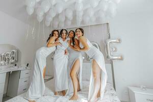 Young bridesmaids in white silk dresses on a bed in the bride's room. Beautiful women celebrating bachelorette party sitting on bed. photo