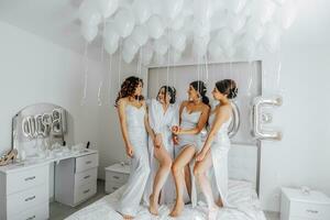 joven damas de honor en blanco seda vestidos en un cama en el de la novia habitación. hermosa mujer celebrando despedida de soltera fiesta sentado en cama. foto