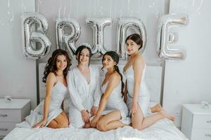 joven damas de honor en blanco seda vestidos en un cama en el de la novia habitación. hermosa mujer celebrando despedida de soltera fiesta sentado en cama. foto