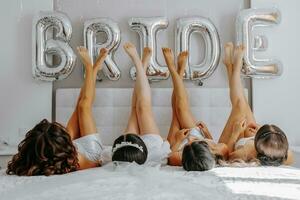 Young bridesmaids in white silk dresses on a bed in the bride's room. Beautiful women celebrating bachelorette party sitting on bed. photo