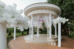 el cerrado Boda arco en el parque es hecho de blanco flores en blanco columnas lejos Boda ceremonia. foto