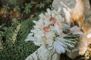 wedding bouquet of pink roses, feathers and dry flowers on the stones in the park photo