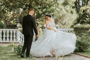 portrait of a young beautiful wedding couple in the garden, the bride in an off-the-shoulder wedding dress circling and showing off her dress, the groom in a black classic suit photo