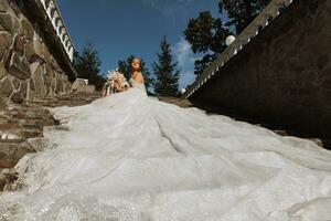 young beautiful bride in a wedding dress with open shoulders and a crown on her head is lying on stone stairs, fashion shot under harsh sunlight photo