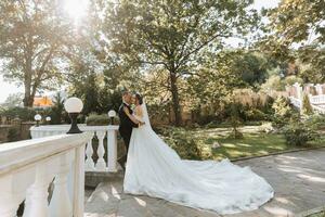 brunette bride in a chic dress with a long train next to the groom in a black classic suit, in a beautiful park with stone steps and white railings photo