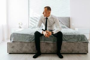 Young handsome businessman sitting on bed in hotel room. The groom is preparing for the wedding. photo