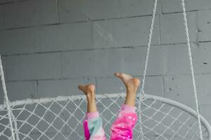 Happy little girl on the porch sitting on a wicker swing and playing in pink pajamas, legs up, close-up photo