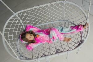 Happy little girl on the porch lying on a wicker swing and playing in pink pajamas with her little toy photo