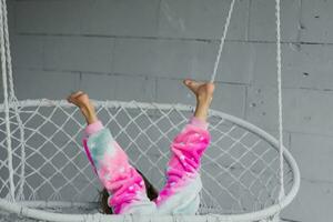 contento pequeño niña en el veranda sentado en un mimbre columpio y jugando en rosado pijama, levanta su piernas a el parte superior foto