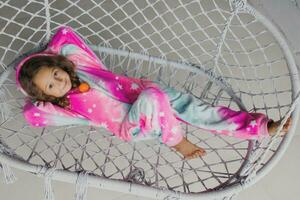 Happy little girl on the porch lying on a wicker swing and playing in pink pajamas with her little toy photo