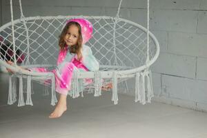 Portrait of happy little girl on porch sitting on wicker swing and playing in pink pajamas photo