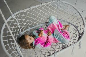 Happy little girl on the porch lying on a wicker swing and playing in pink pajamas with her little toy photo