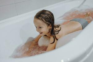 Small, smiling, beautiful dark-haired girl with long hair, child bathes, washes in a white bath with foam. Fun photo. photo