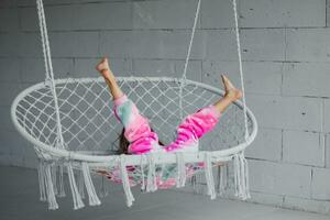 Happy little girl on the veranda sitting on a wicker swing and playing in pink pajamas, raises her legs to the top photo