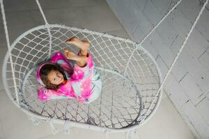 A happy little girl in pink pajamas is lying on a wicker swing and playing with her legs up on the porch. photo