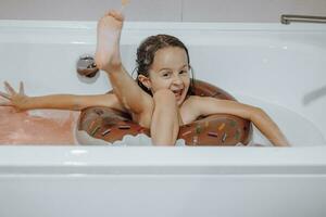 Small, smiling, beautiful dark-haired girl with long hair, child bathes, washes in a white bath with foam. Fun photo. photo