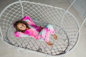 Happy little girl on the porch lying on a wicker swing and playing in pink pajamas with her little toy photo