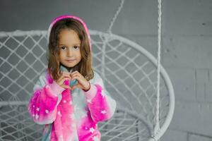 Happy little girl sitting on a wicker swing on the porch and playing in pink pajamas and making a heart shape with her hands. Children's friendship. photo