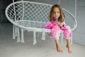 retrato de un contento pequeño niña en rosado pijama en el veranda, acostado en un mimbre columpio y jugando con su pequeño juguete foto