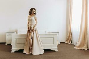 A beautiful young female model in a stylish evening dress made of light satin fabric poses in a white room. photo