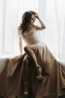 A happy beautiful girl with closed eyes elegantly sits on the windowsill. A princess with long flowing hair in a luxurious beige dress. Graduation party photo