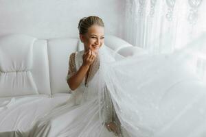 A gorgeous bride with a classic hairstyle, wearing a long robe and a long veil, is sitting and posing on a white sofa in her room. The bride on a white background photo