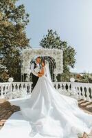 Stylish groom in white shirt and black vest and cute blonde bride in white dress in park near elegant white rose arch. Wedding portrait of newlyweds. photo