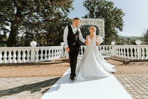 elegante novio en blanco camisa y negro chaleco y linda rubia novia en blanco vestir en parque cerca elegante blanco Rosa arco. Boda retrato de recién casados. foto