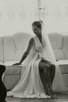 Black and white photo. A gorgeous bride with a classic hairstyle, wearing a long robe and a long veil, is sitting and posing on a white sofa in her room. The bride on a white background. photo