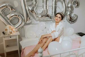 A beautiful brunette bride in a white robe, a veil and a tiara on her head, sits on a bed. Inscription bride. Happy young girl under the glow of sunlight. wedding concept photo