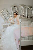 A beautiful young bride in a wedding dress with a bouquet of peonies in her hands is sitting on a bed in a room. Final preparations for the wedding. The bride is waiting for the groom. Morning, bride. photo