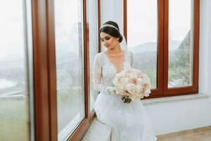 the bride in an evening dress sits on the windowsill against the background of large windows. Beautiful young bride. Free space photo