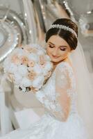 A beautiful young bride in a wedding dress with a bouquet of peonies in her hands is sitting on a bed in a room. Final preparations for the wedding. The bride is waiting for the groom. Morning, bride. photo