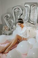 Brunette bride in white robe and veil, sitting on bed, posing and wearing leg bandage, bare legs. Wedding concept. Morning of the bride. Classic hair and makeup photo