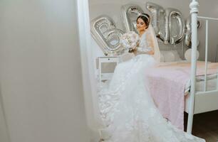 A beautiful young bride in a wedding dress with a bouquet of peonies in her hands is sitting on a bed in a room. Final preparations for the wedding. The bride is waiting for the groom. Morning, bride. photo