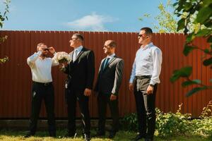 The groom in a black suit and glasses and his stylish friends wearing white shirts and black pants and glasses are standing in the backyard in the garden. The groom is holding a bouquet. photo