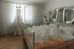 A lace dress embroidered with flowers with a train hangs on the eaves near the window in the bride's room. A lot of white balls on the bed, the inscription bride with silver balls. photo