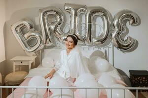 un hermosa morena novia en un blanco túnica, un velo y un tiara en su cabeza, se sienta en un cama. inscripción novia. contento joven niña debajo el resplandor de luz de sol. Boda concepto foto