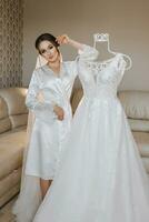 A beautiful woman with hair on her head, make-up and a wedding veil in a dressing gown is standing next to her dress on a mannequin. Dressing up and preparing for the wedding ceremony photo