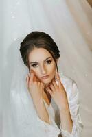 Pretty woman with hairstyle, make-up and wedding veil sitting in dressing gown on sofa, dressing and preparing for wedding ceremony photo