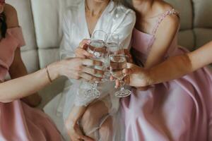 the bride, with girlfriends in identical pink dresses, in the morning, sitting on the sofa, celebrate and rejoice with glasses of champagne in their hands photo