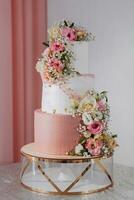Three-tiered white wedding cake decorated with various flowers on silver surface and pink background in wedding hall. photo