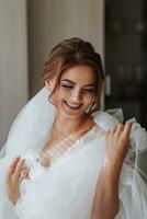 A bride with brown hair in a robe, posing for the camera, looking at her wedding dress. Elegant hairstyle. Nice makeup. Voluminous veil. Morning of the bride photo