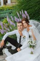 Portrait of the bride and groom in the park. The bride is holding a bouquet and hugging her groom, they are sitting, posing near lavender flowers. Wedding walk in the park. Long train of the dress photo