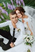 Portrait of the bride and groom in the park. The bride is holding a bouquet and hugging her groom, they are sitting, posing near lavender flowers. Wedding walk in the park. Long train of the dress photo