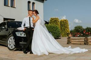 gorgeous bride with trendy makeup and hairdo in luxury wedding dress with handsome groom near black wedding car photo